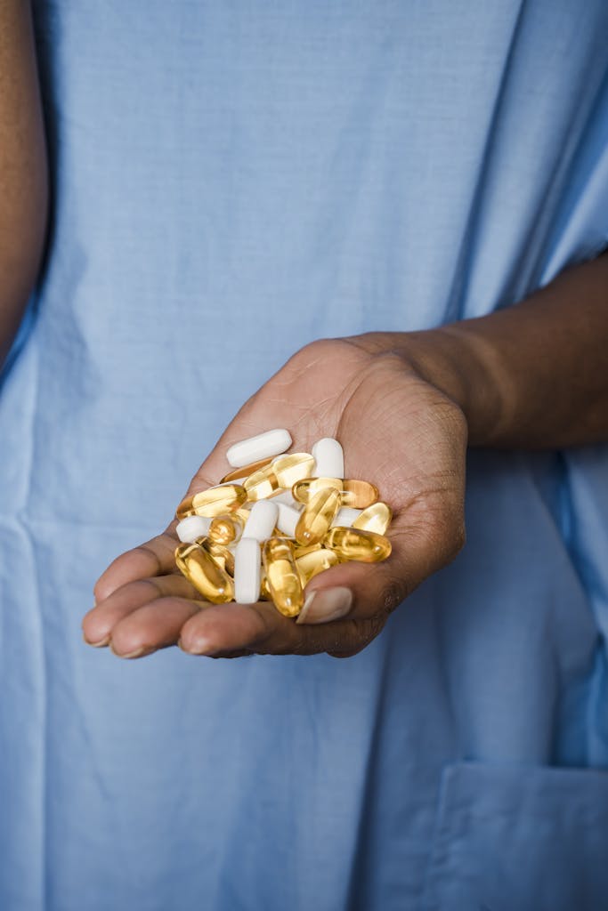 Crop unrecognizable black patient in blue medical robe demonstrating white and yellow medical pills in hand in bright room
