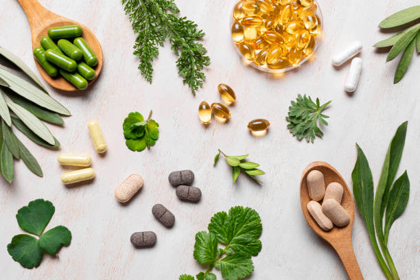 Natural vitamins, plant based supplements and green leaves flat lay on white wooden background. Various organic capsules and pills.
